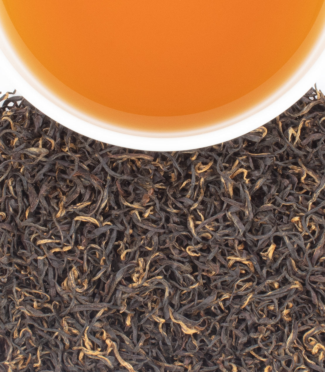 A cup of amber-colored tea rests above a pile of dark, curly loose-leaf tea.