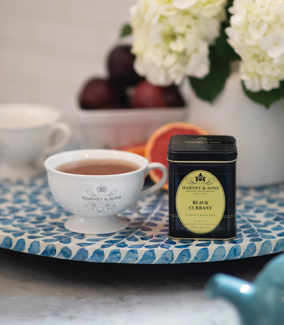 Black Currant Tea by Harney & Sons in a tin, with a cup of brewed tea and fresh fruit on a decorative tray.