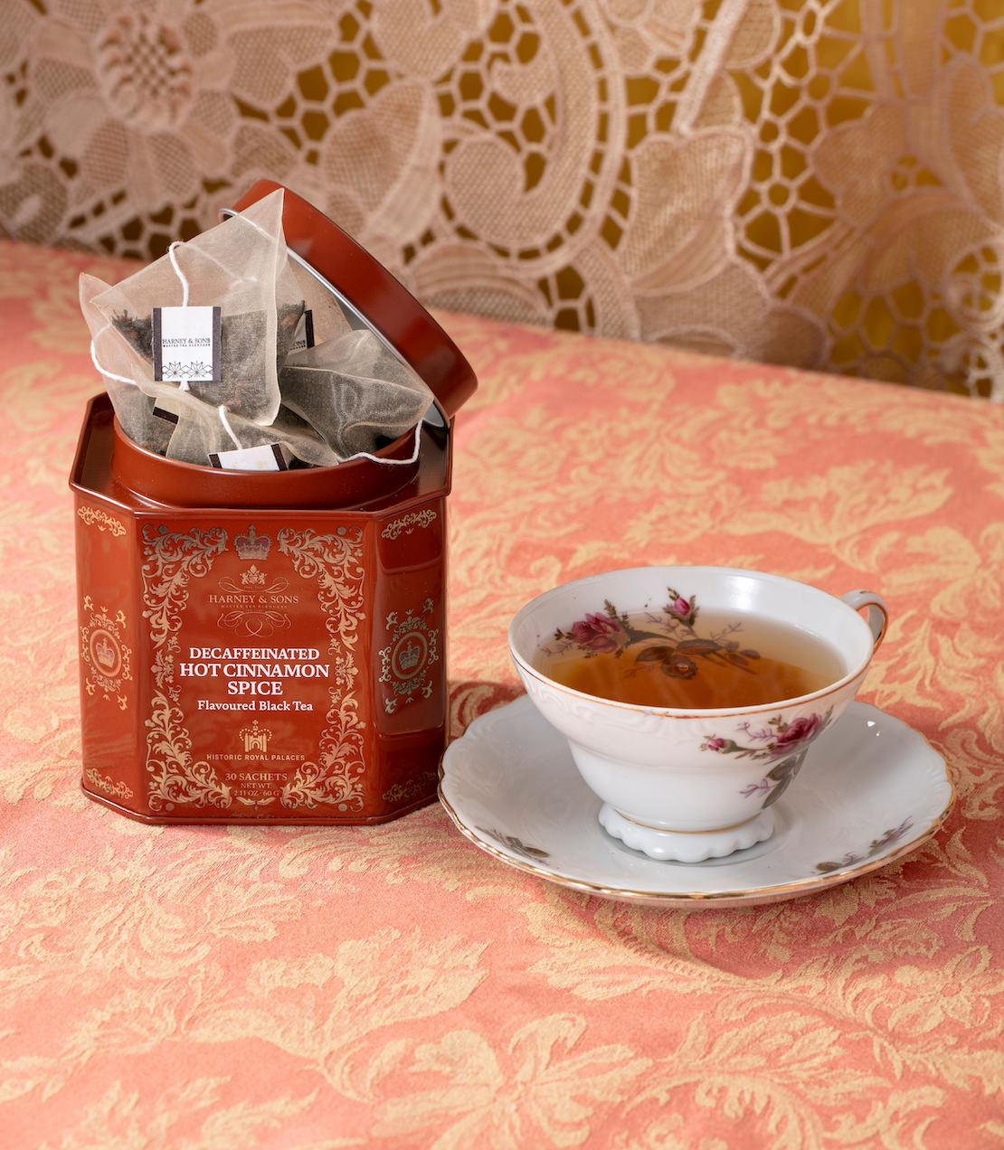 Tea tin with sachets and a cup of tea on a patterned tablecloth.