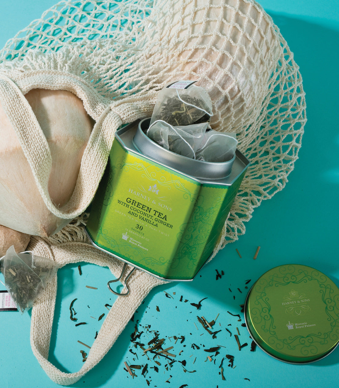 Green tea tin with coconut, ginger, and vanilla sachets, placed in a natural bag on a turquoise background.