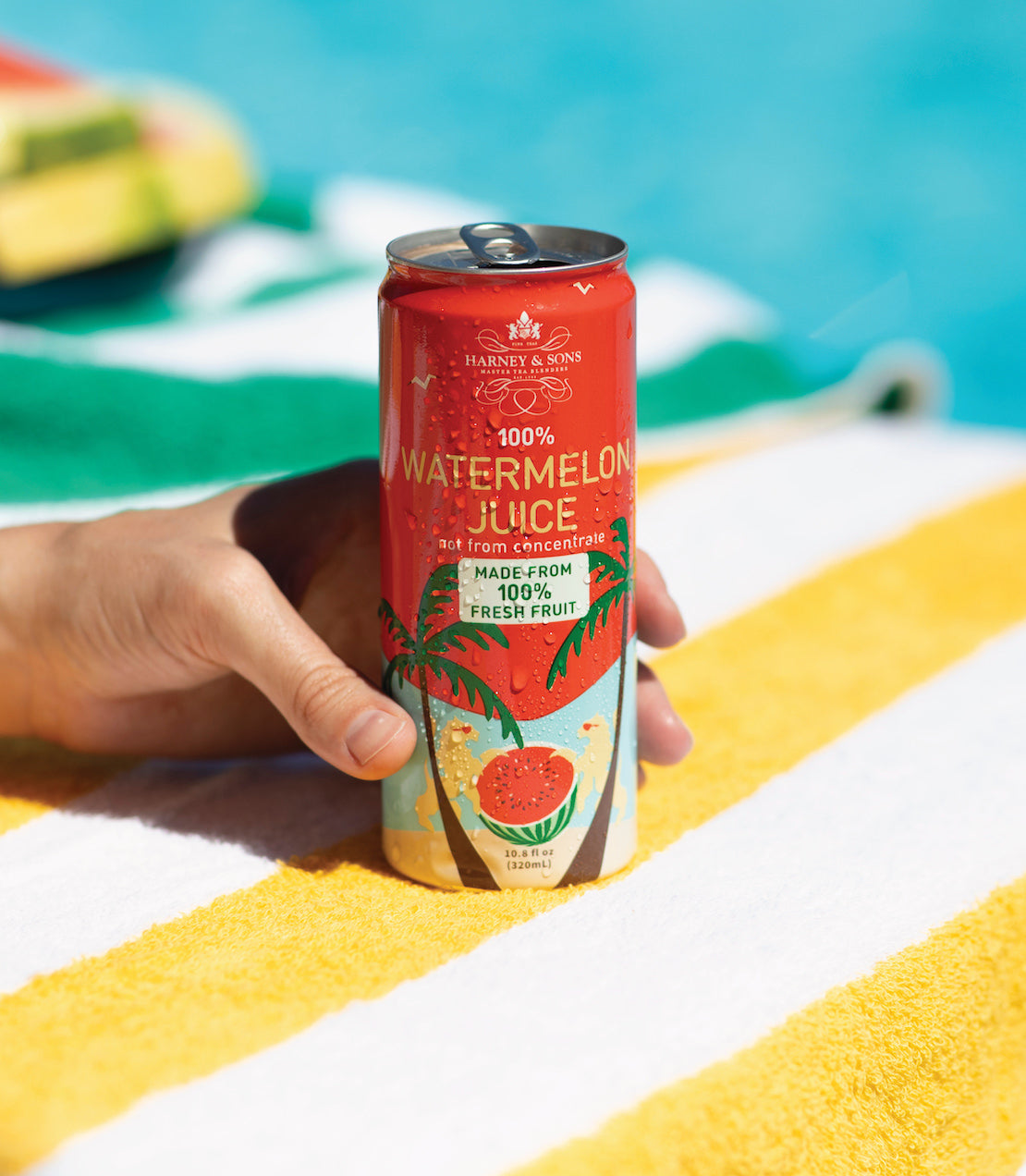 Hand holding a can of Harney & Sons 100% Watermelon Juice, featuring vibrant tropical design, on a striped beach towel.