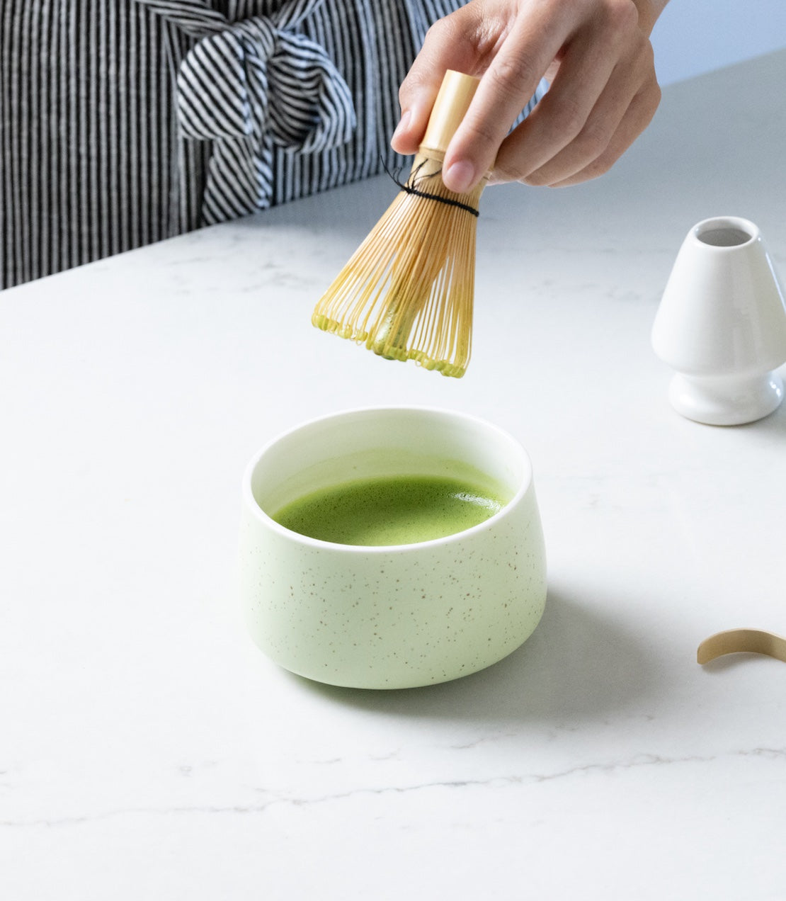 Person preparing matcha tea with a whisk and a green bowl on a white surface.