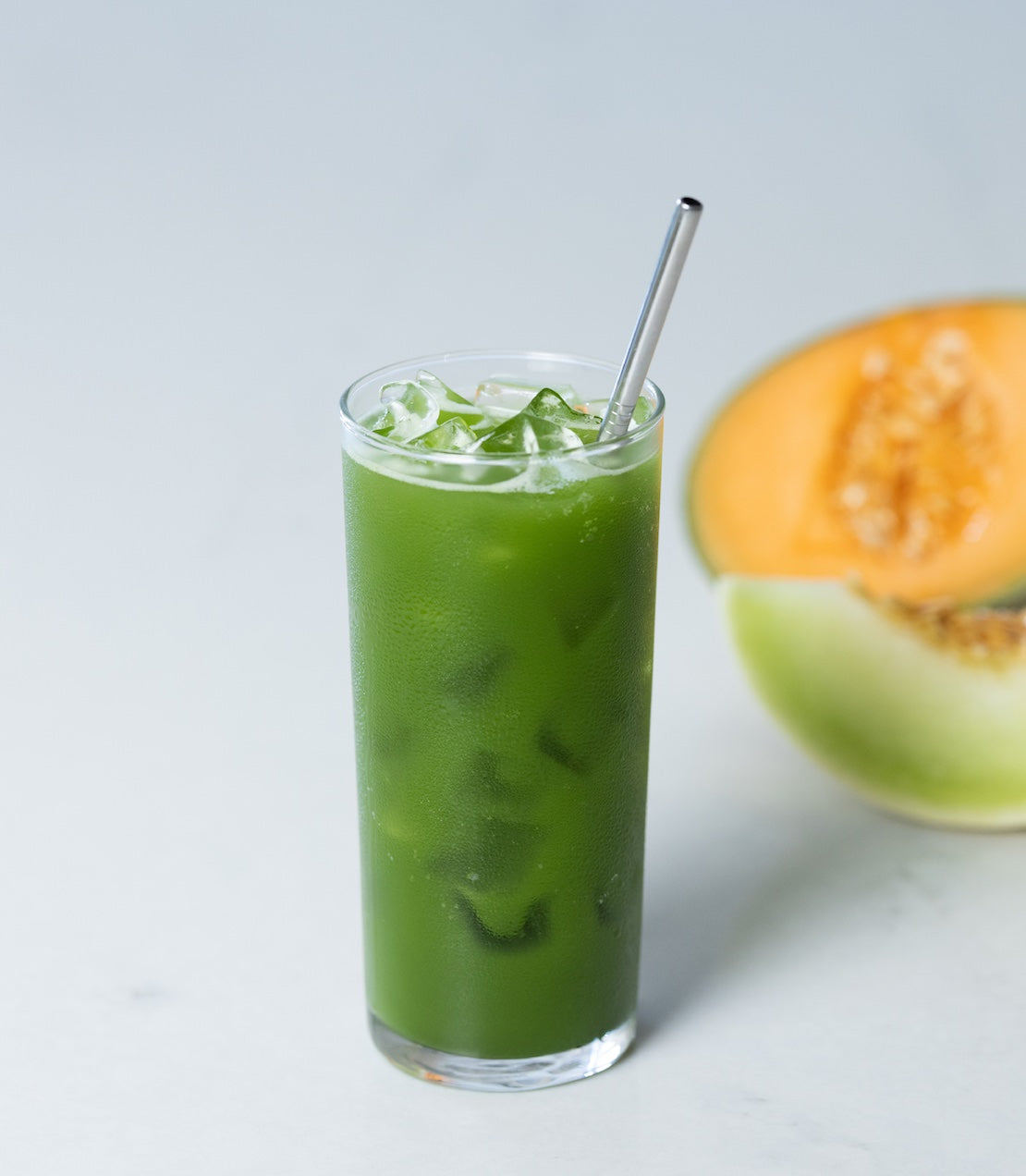 Green iced matcha drink in a tall glass with a metal straw, next to a halved cantaloupe on a light background.