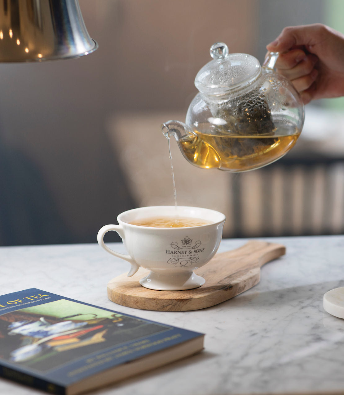 Pouring Milky Oolong tea from a glass teapot into a white cup, with a book on a wooden board nearby.