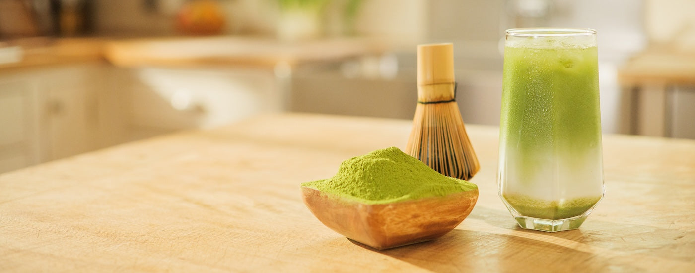 A glass of green matcha tea on a wooden table next to a small wooden bowl filled with matcha powder and a bamboo whisk, with a softly blurred kitchen background.