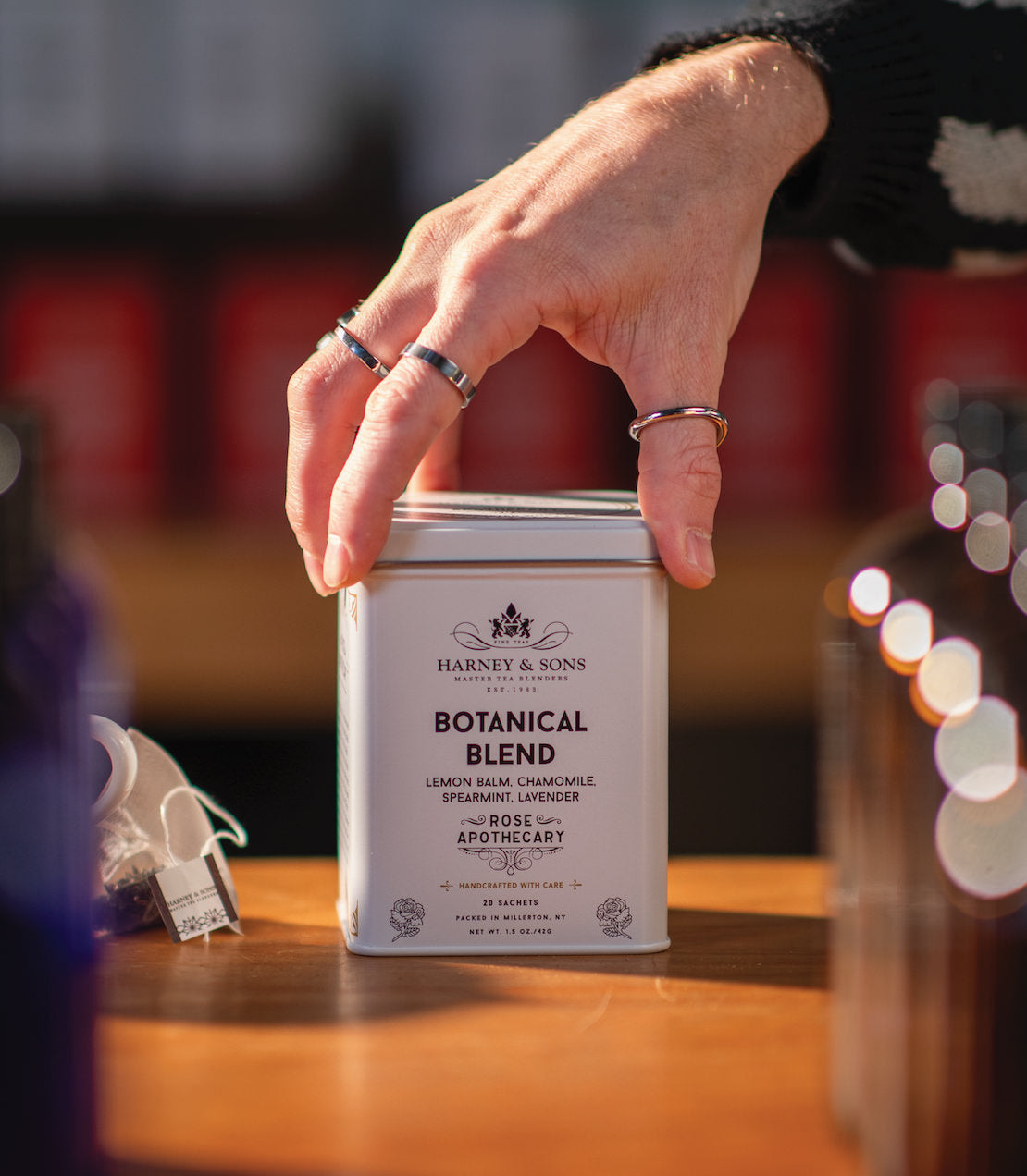 Hand reaching for a tin of Harney & Sons Botanical Blend tea sachets, featuring lemon balm, chamomile, and lavender.
