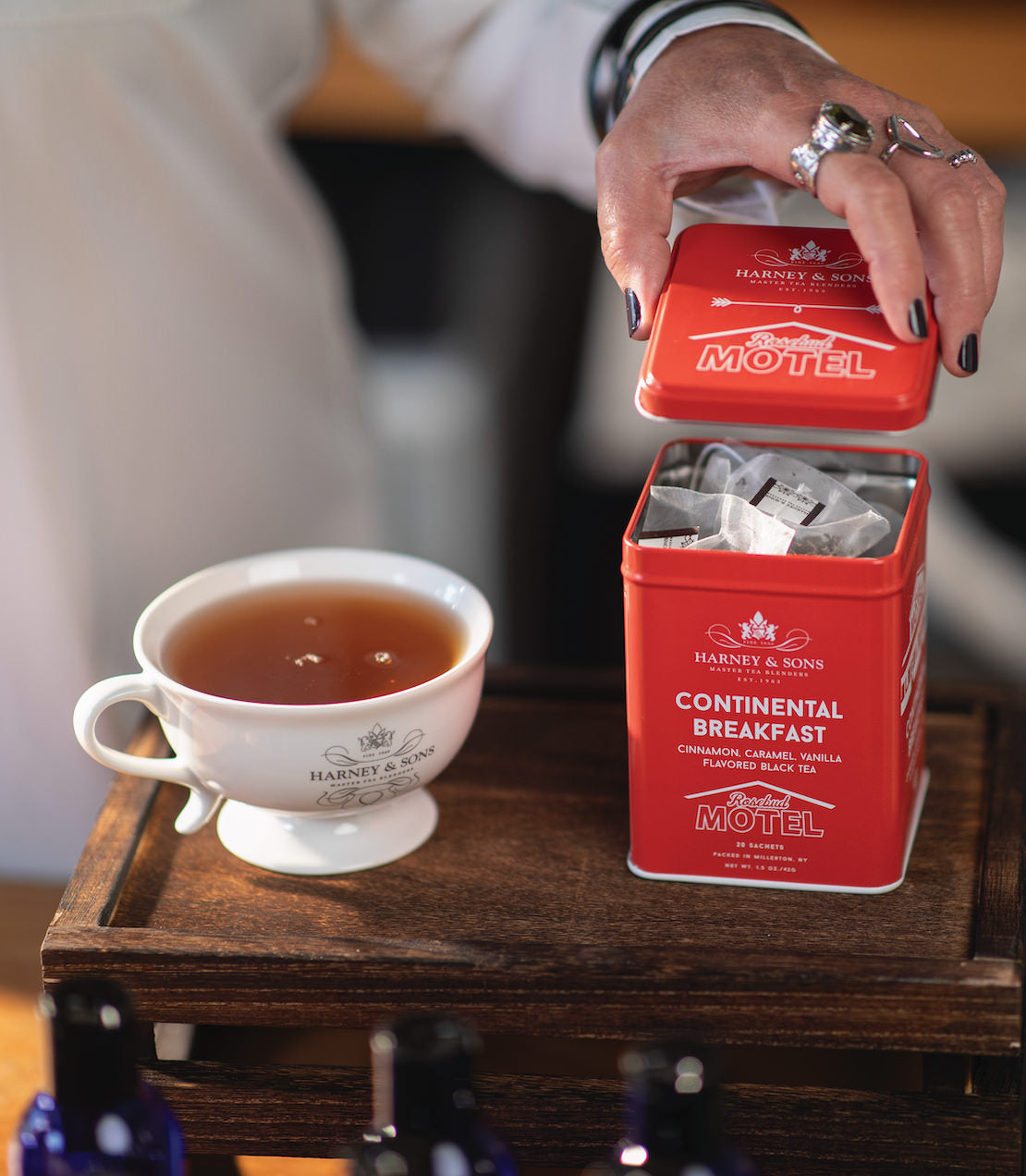 Hand pouring sachets from a red tin labeled "Continental Breakfast" into a cup of tea on a wooden tray.