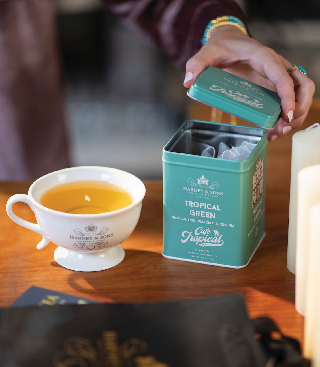 Hand opening a tin of Tropical Green tea sachets beside a cup of brewed tea on a wooden table.