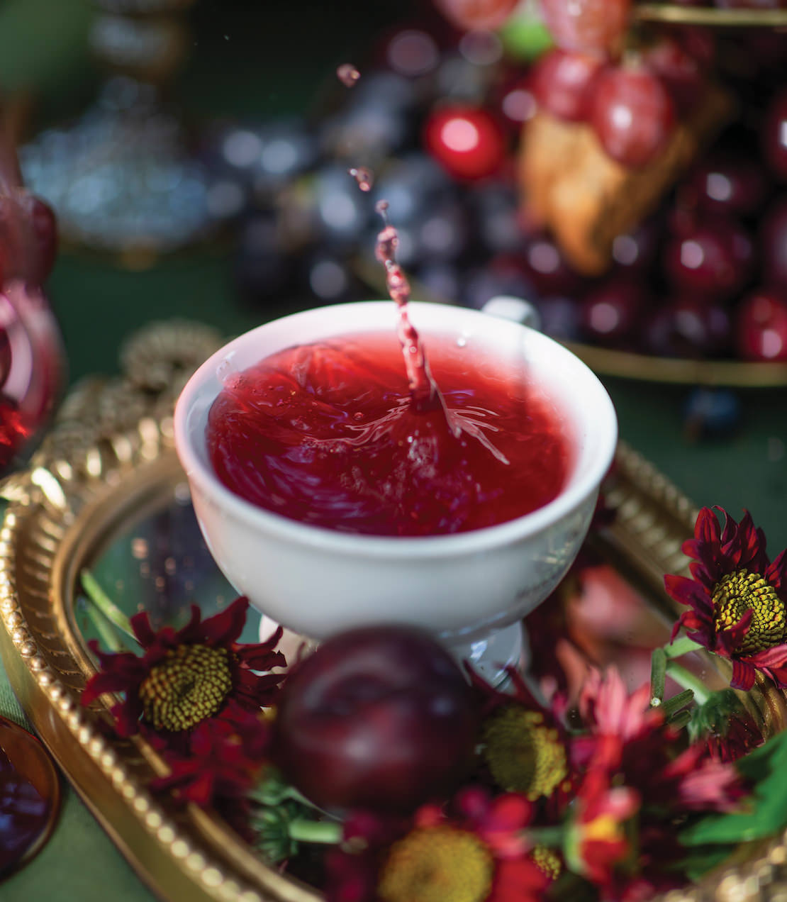 A cup of spiced plum herbal tea with a splash, surrounded by fresh plums and vibrant flowers on a decorative tray.