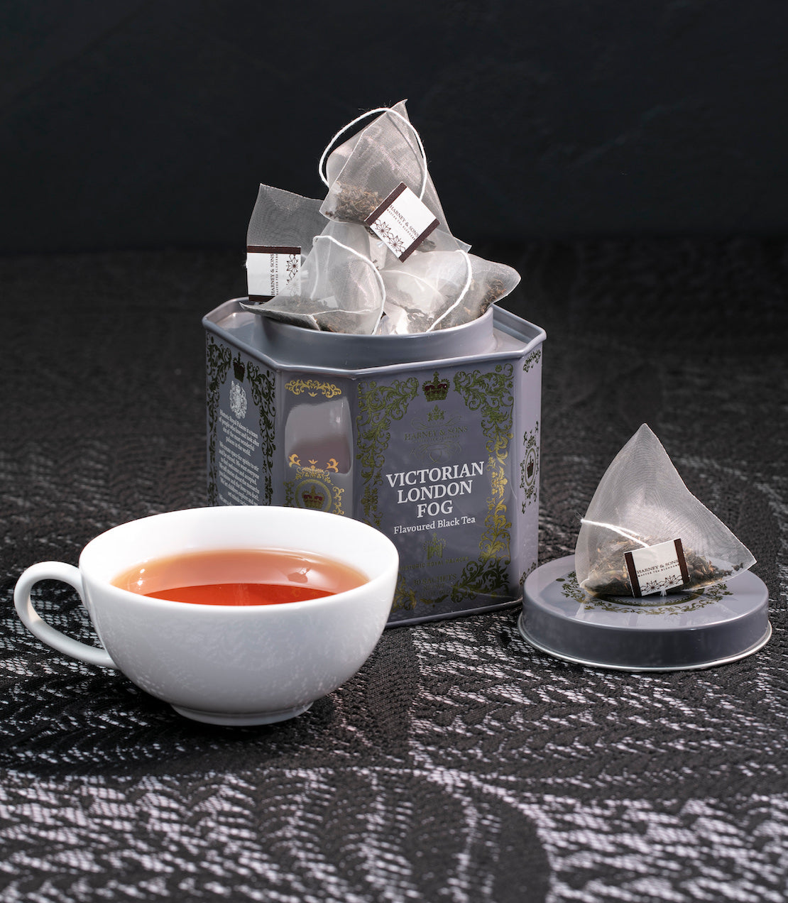 Tea tin with a cup of tea and sachets on a dark surface.