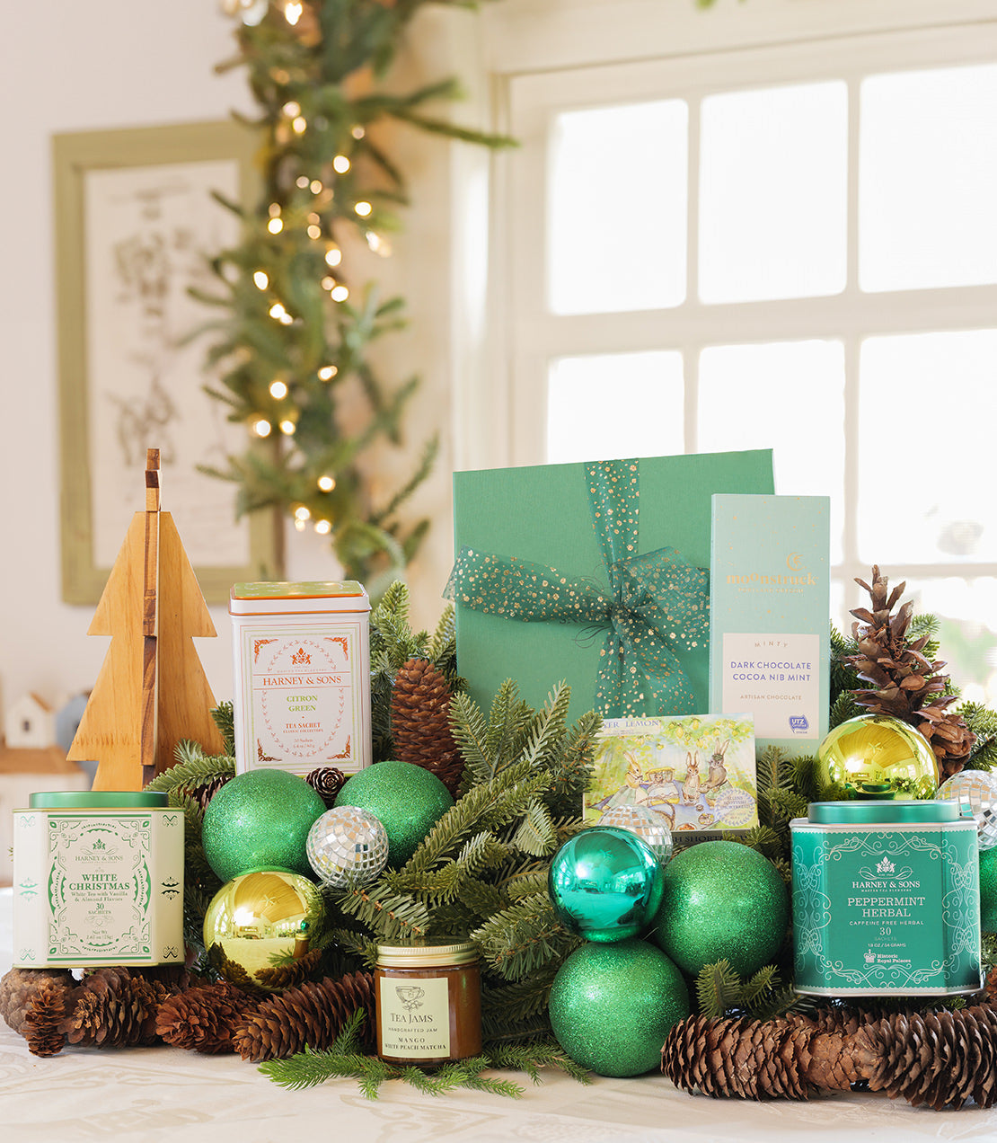 Festive holiday gift arrangement featuring green ornaments, candles, chocolates, and a decorative tree.
