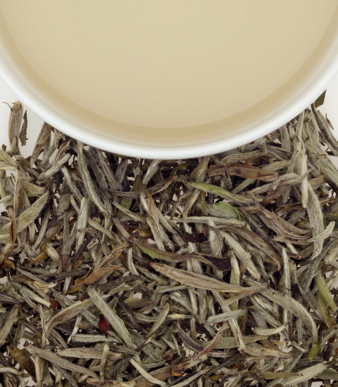 Close-up of Chinese Silver Needle tea leaves beside a cup of pale yellow tea.