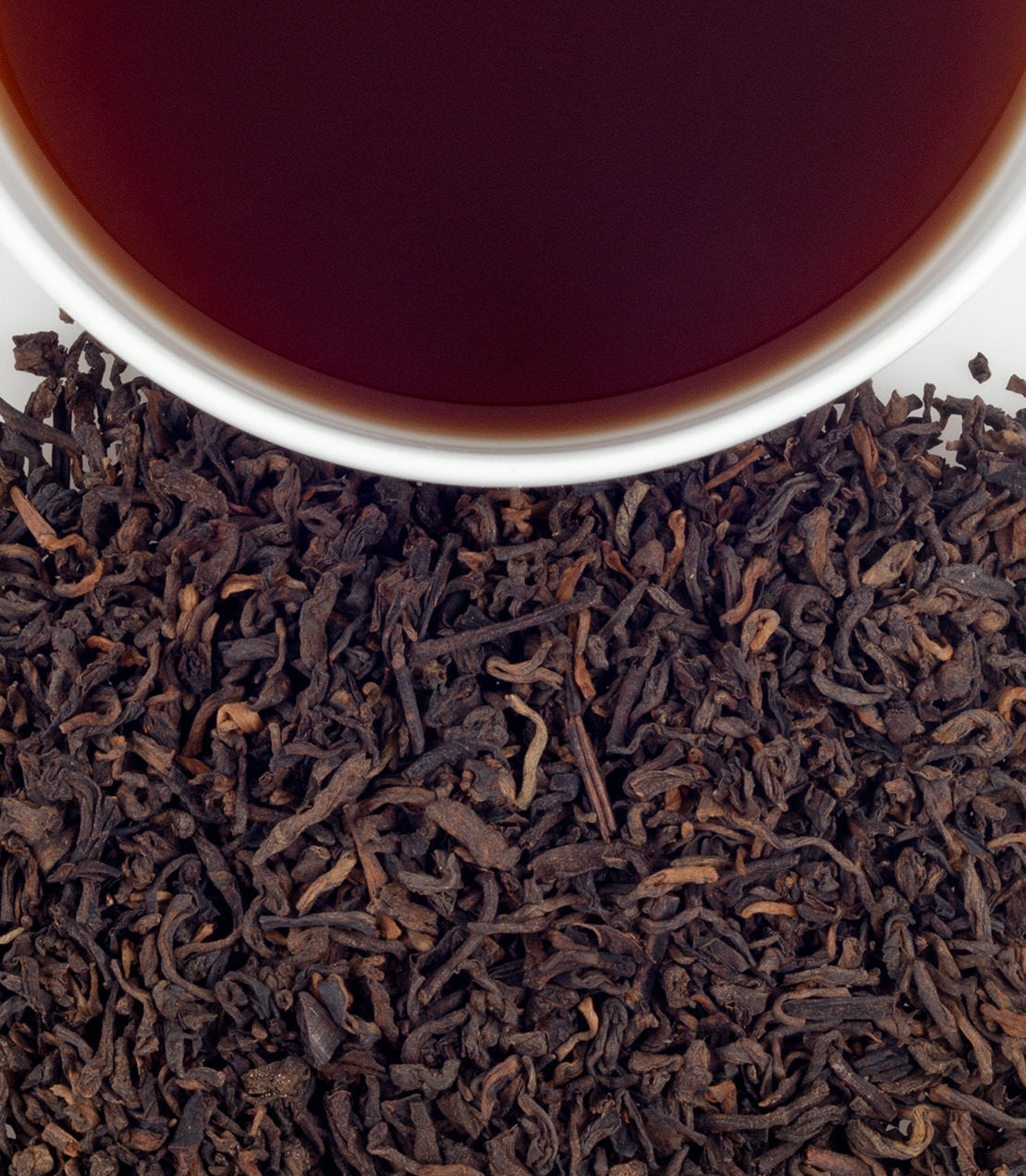 A close-up of loose Pu-Erh tea leaves with a cup of brewed tea above, showcasing its rich, dark color.