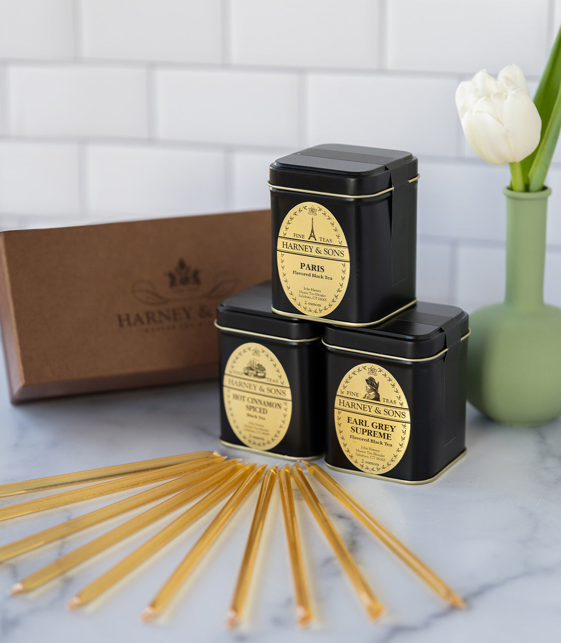 Three black tea tins from Harney & Sons, including Paris and Earl Grey Supreme, with honey sticks and a vase.